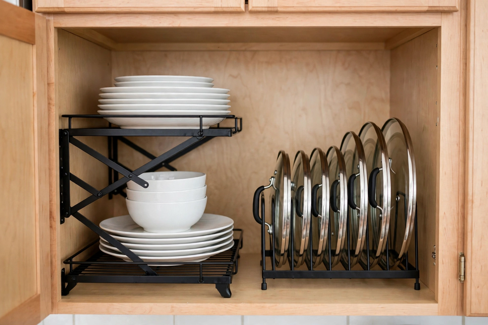 Expandable black metal shelf riser holding stacked plates and bowls, with a pot lid organizer rack holding lids upright inside a kitchen cabinet